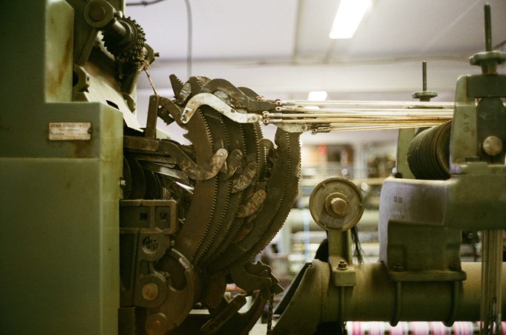 Side view of a loom in a European weaving mill, showing the shedding mechanism that lifts warp threads to create space for the weft. The interplay of levers and cams controls the rhythm of the loom, while the warp threads are held under tension from the beam.